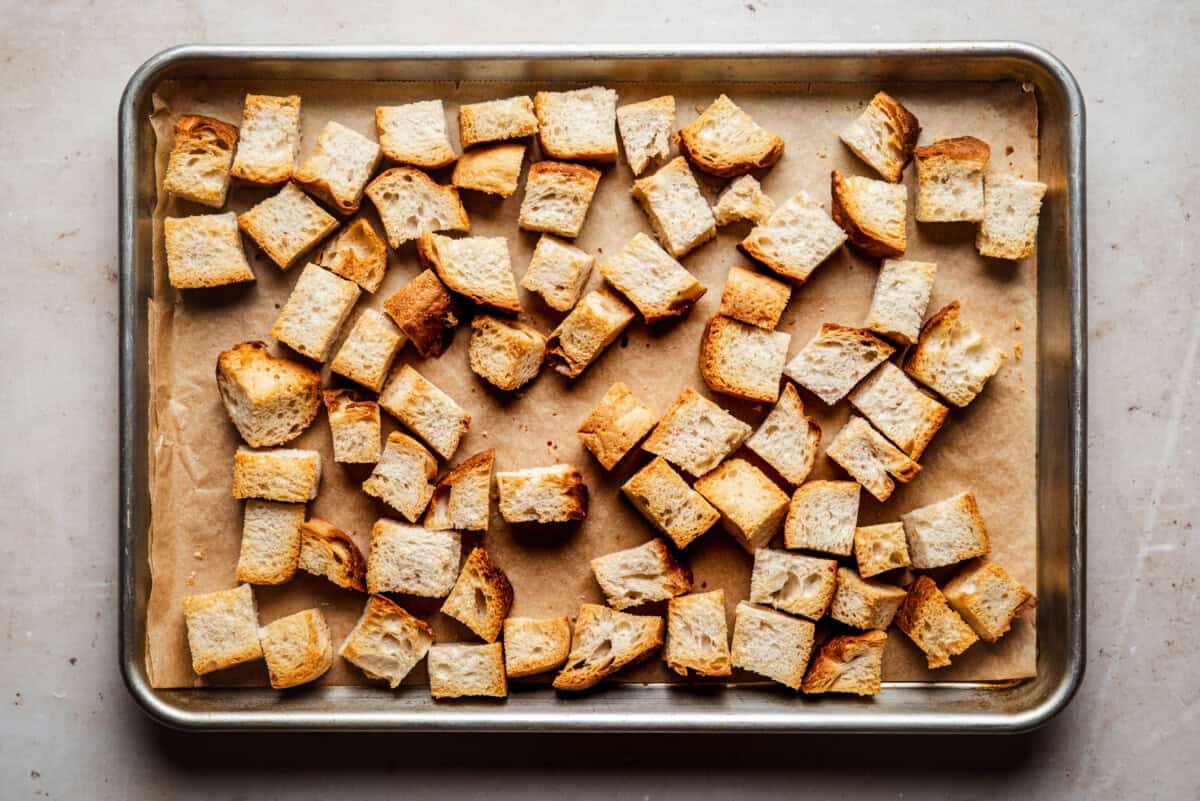 Cubed pieces of toasted bread are spread out on a parchment-lined baking sheet, ready to be used as croutons.