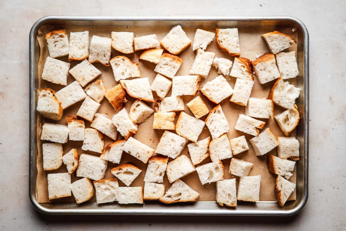 A baking tray lined with parchment paper holds evenly spread cubes of sliced bread, ready for toasting or baking.
