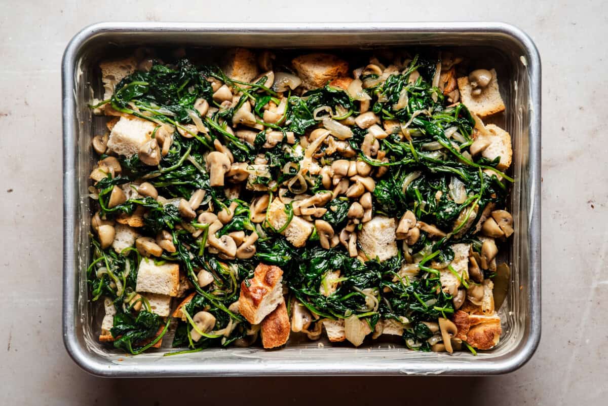 A baking dish filled with sautéed spinach, mushrooms, onions, and cubes of toasted bread, arranged evenly and ready to be baked.