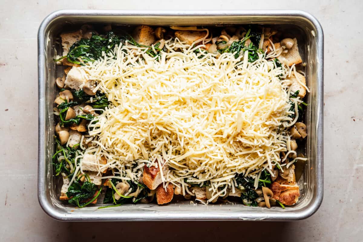 A rectangular metal baking dish filled with a mixture of sautéed spinach, mushrooms, bread cubes, and topped with a generous layer of shredded cheese, ready to be baked.