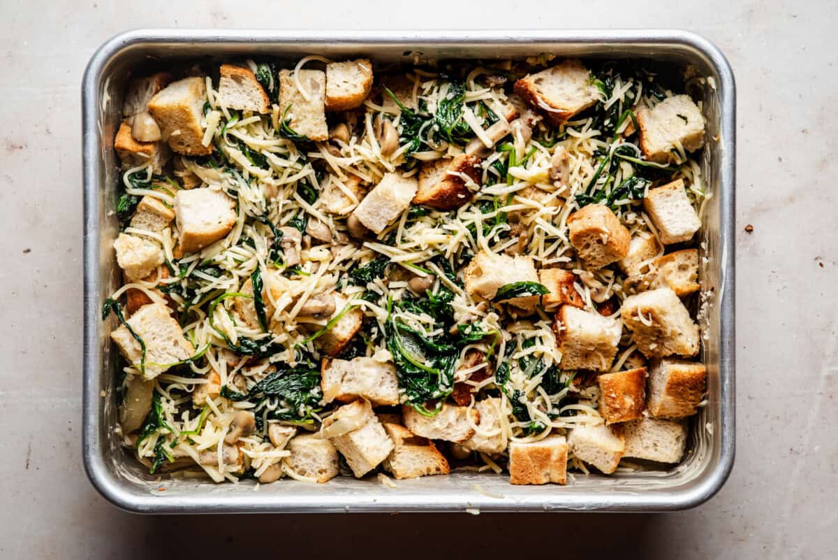 A metal baking dish filled with a casserole made of bread cubes, wilted spinach, shredded cheese, and sautéed vegetables, ready to be baked.