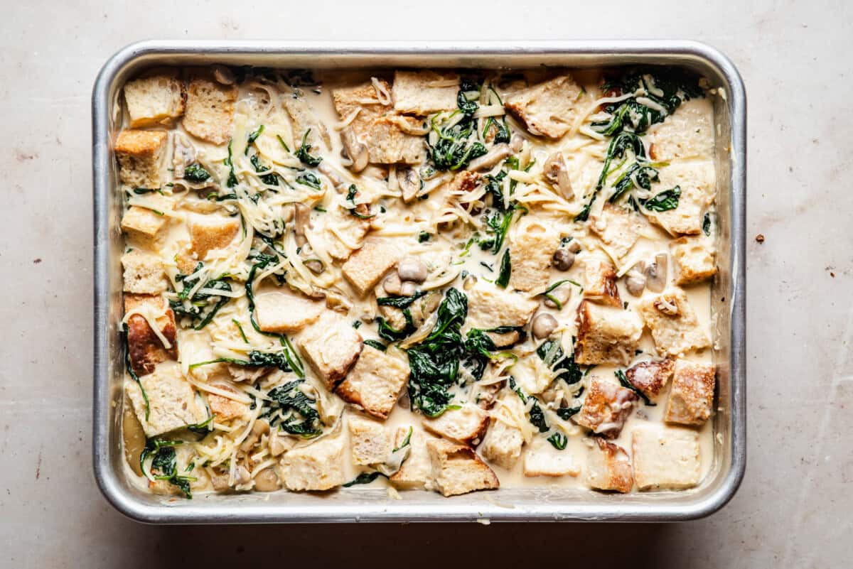 A baking dish filled with an unbaked casserole made of cubed bread, spinach, mushrooms, shredded cheese, and a creamy sauce on a light countertop.