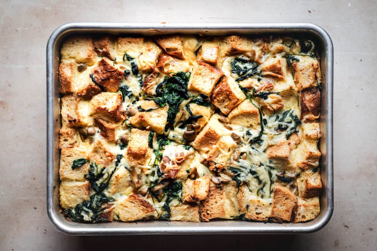 A baked bread pudding in a rectangular metal pan, featuring golden bread cubes, wilted greens, and a creamy mixture on a light countertop.