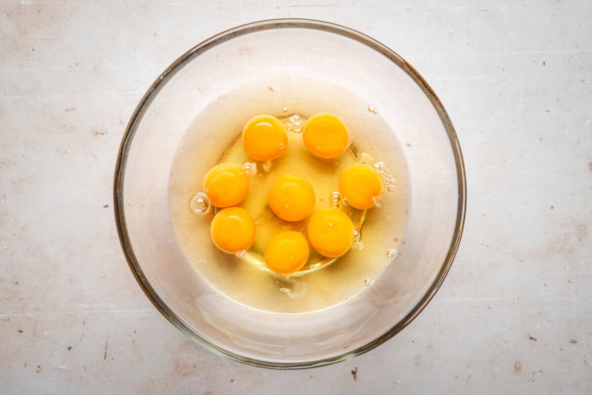 A glass bowl containing seven raw eggs with unbroken yolks, placed on a light-colored surface.