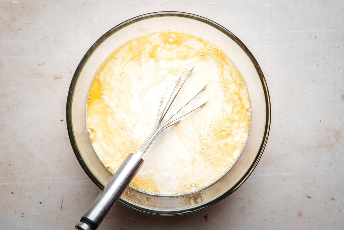 A whisk rests in a glass bowl filled with a mixture of eggs and milk, sitting on a light-colored countertop.