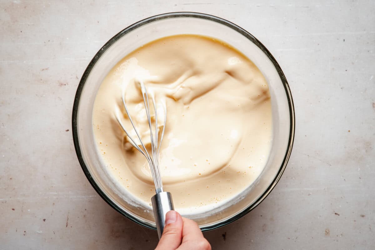 A hand holding a metal whisk is mixing a thick, creamy batter in a clear glass bowl on a light-colored surface.