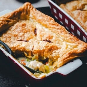 Golden, flaky puff pastry tops a rectangular baking dish filled with creamy turkey pot pie, visible chunks of turkey, carrots, and celery; a serving spoon rests inside the dish.