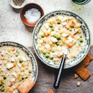 Two bowls of creamy chicken and vegetable soup with peas, carrots, and potatoes, garnished with herbs. A black-handled spoon rests in one bowl. Crackers, salt, and spices are placed nearby on a rustic surface.