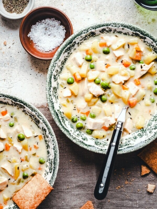 Two bowls of creamy chicken and vegetable soup with peas, carrots, and potatoes, garnished with herbs. A black-handled spoon rests in one bowl. Crackers, salt, and spices are placed nearby on a rustic surface.