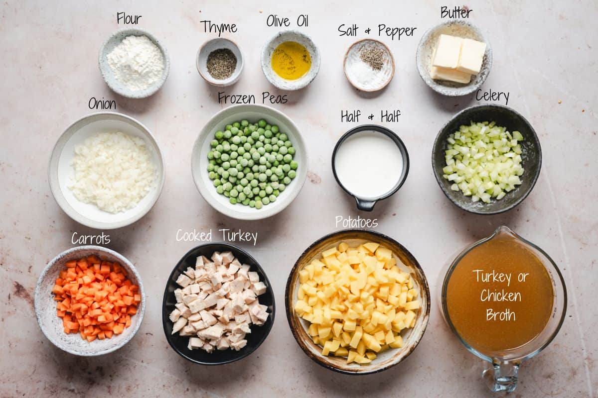 Top-down view of labeled bowls containing ingredients for a turkey pot pie soup recipe: flour, thyme, olive oil, salt and pepper, celery, butter, onion, frozen peas, half & half, carrots, cooked turkey, potatoes, and turkey or chicken broth.