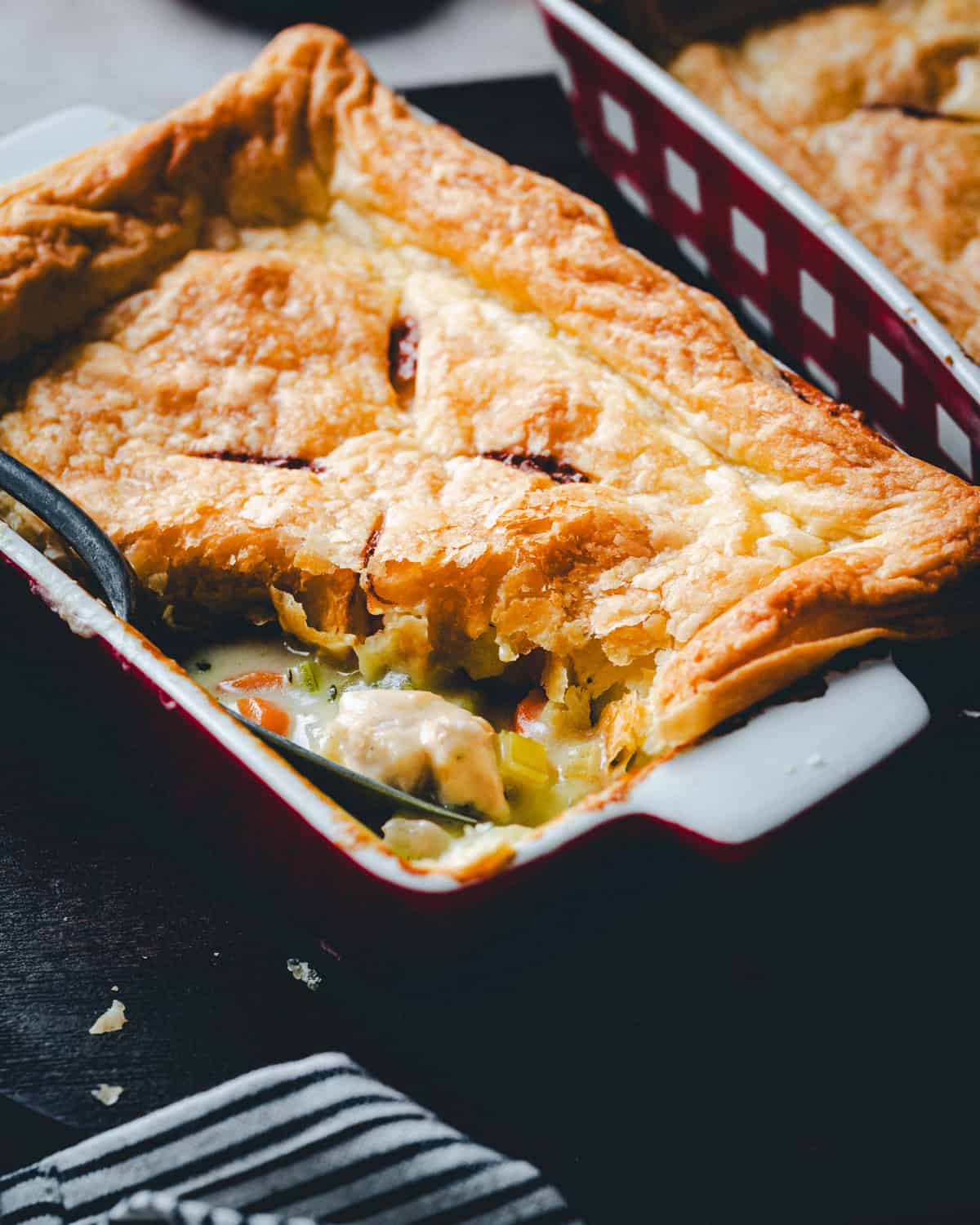 A close-up of a flaky, golden-brown turkey pot pie with a savory filling of vegetables and turkey, served in a red and white baking dish with a spoon scooping out a portion.