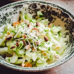 A bowl with a green and white floral pattern filled with a salad of sliced celery, green onions, chopped almonds, and shredded cheese, garnished with fresh herbs.