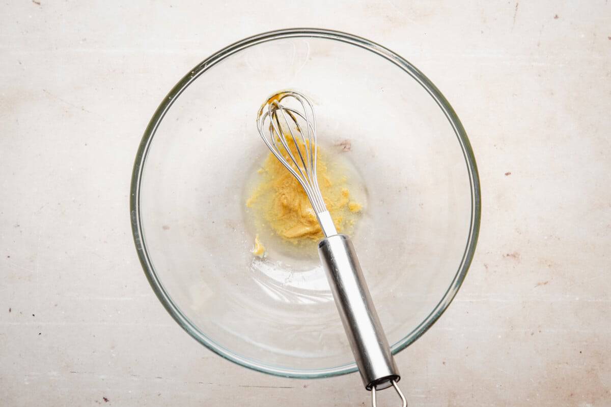 A metal whisk rests in a glass bowl containing a small amount of mustard, honey and lemon juice on a light-colored surface.