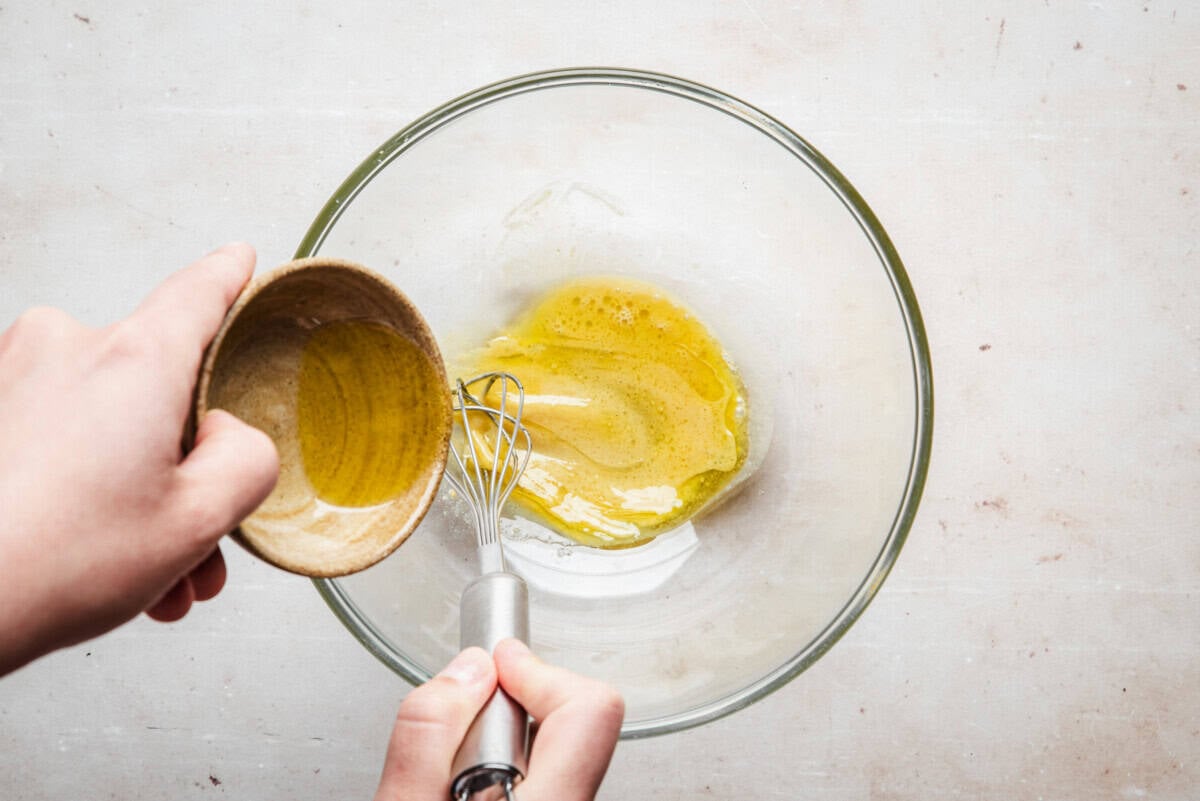 A hand pours oil from a small bowl into a larger glass bowl containing whisked eggs while holding a metal whisk, preparing to mix the ingredients.