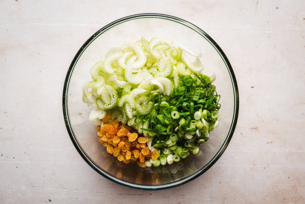 A glass bowl containing sliced celery, chopped green onions, and golden raisins, arranged in sections on a light-colored surface.