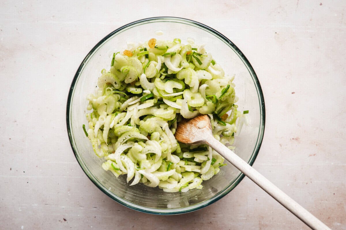 A glass bowl filled with a salad made of sliced celery, onions, and herbs sits on a light surface. A wooden spoon rests in the bowl, mixing the fresh, green ingredients.