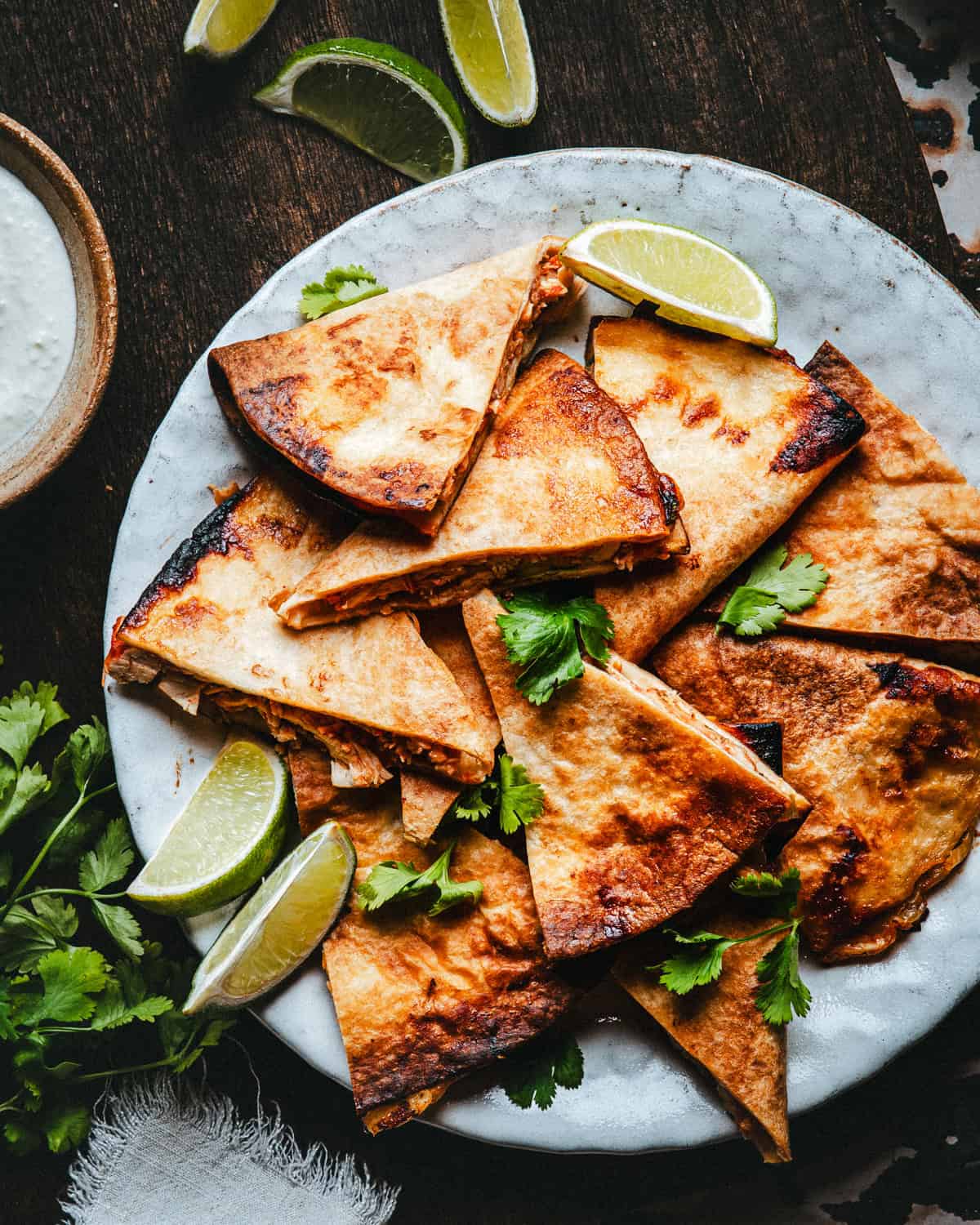 A plate of golden-brown quesadilla wedges garnished with cilantro and lime wedges, placed on a dark wooden surface. A bowl of dipping sauce and additional cilantro are on the side.