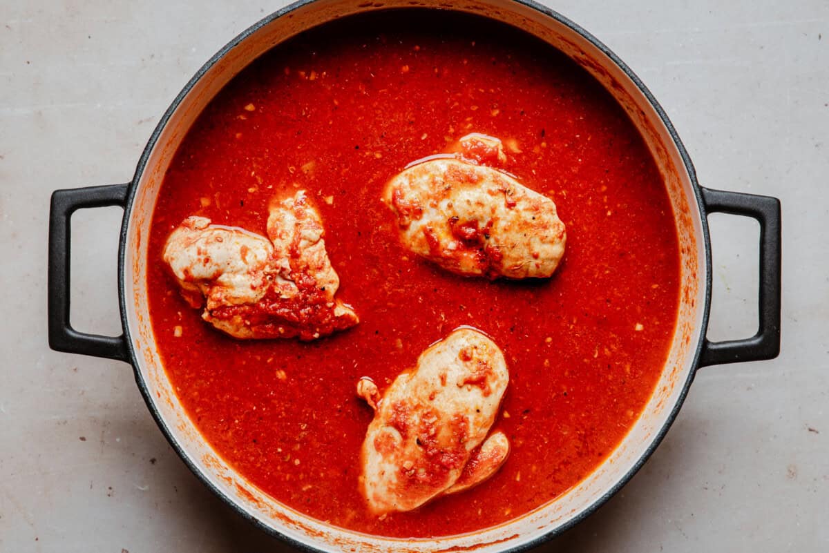 A pot with three pieces of chicken simmering in a thick, red tomato-based sauce, viewed from above on a light-colored surface.