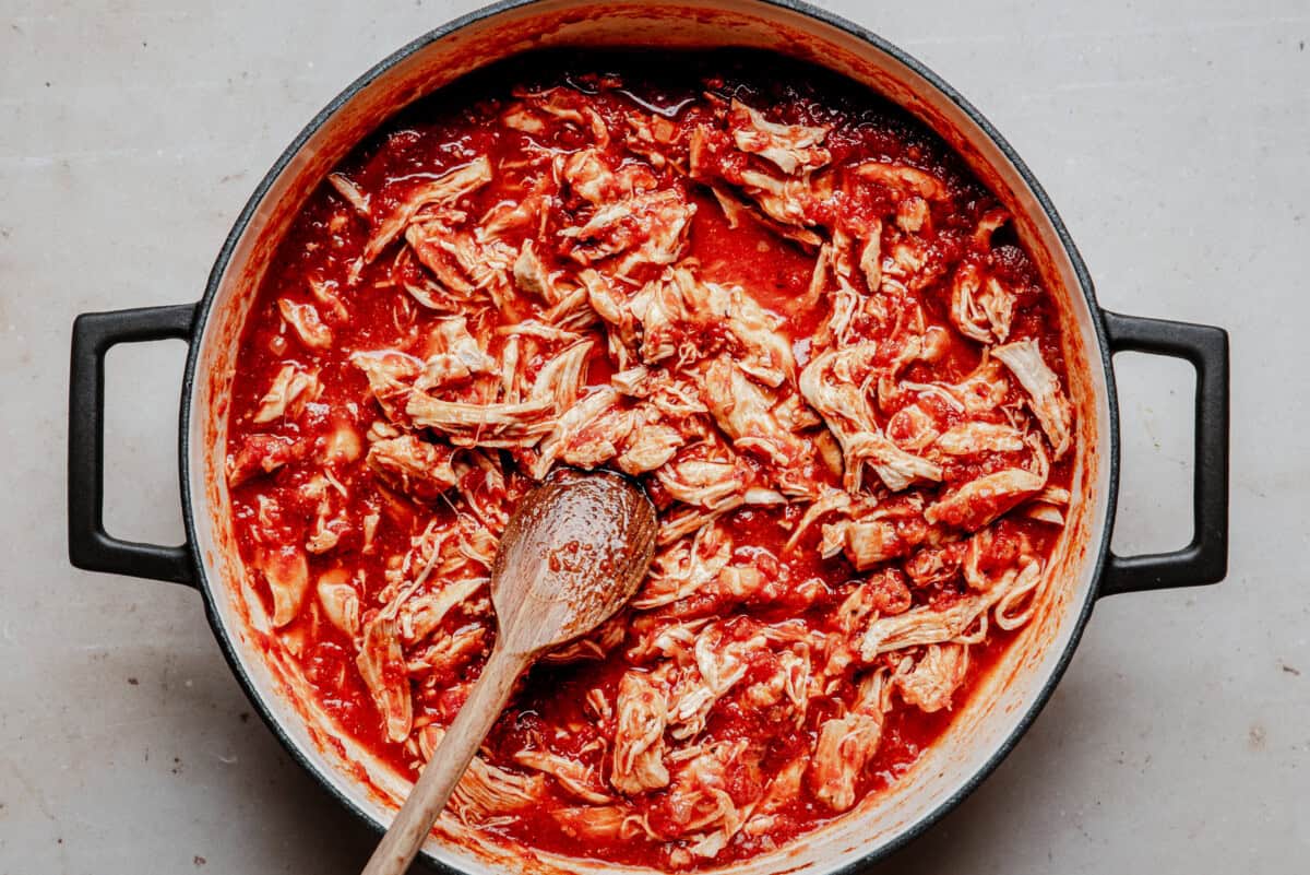 A large pot filled with shredded chicken cooked in a rich red tomato sauce, with a wooden spoon resting inside and sauce spread around the edges.