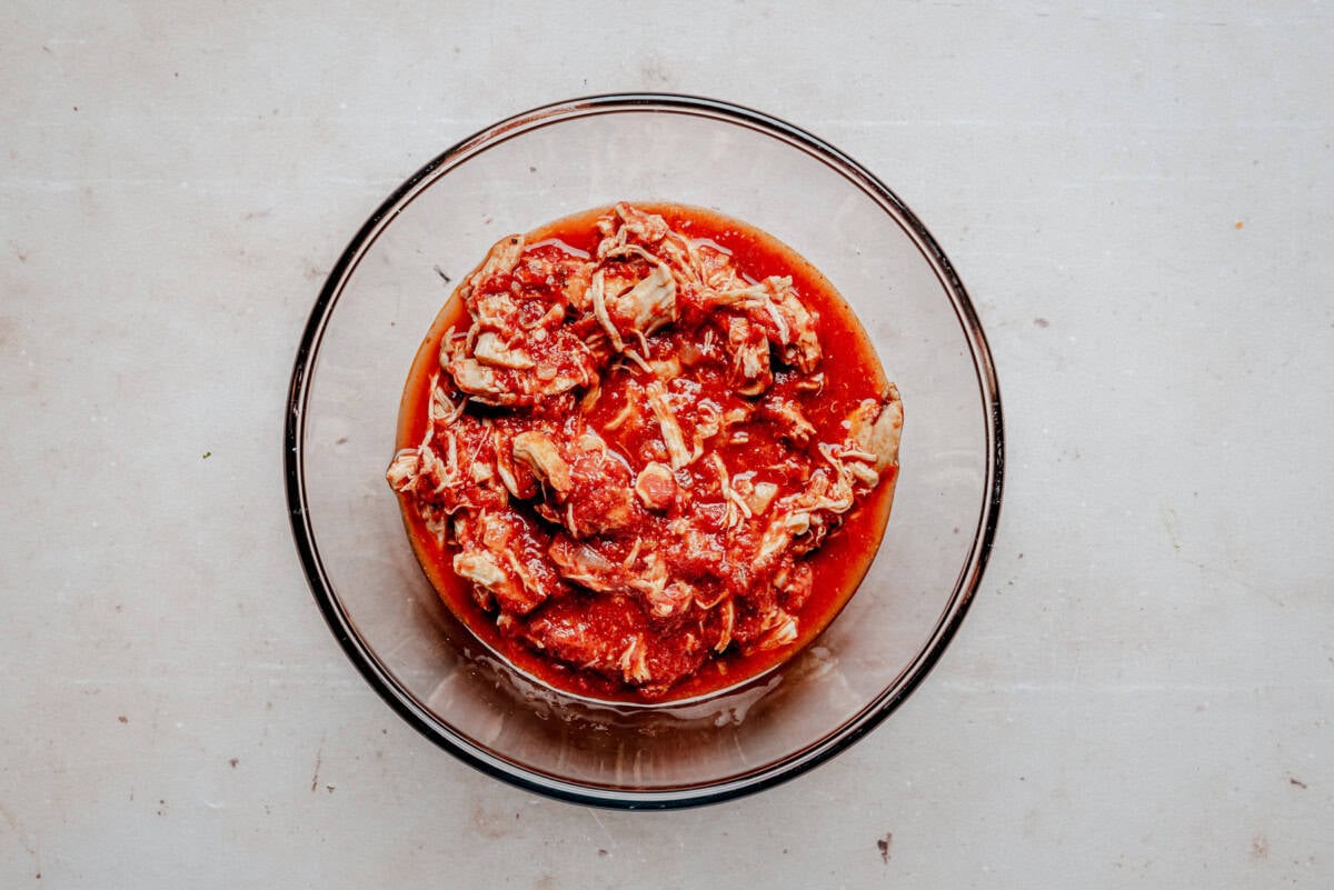A clear glass bowl filled with shredded chicken in a red tomato-based sauce sits on a light-colored, textured surface.