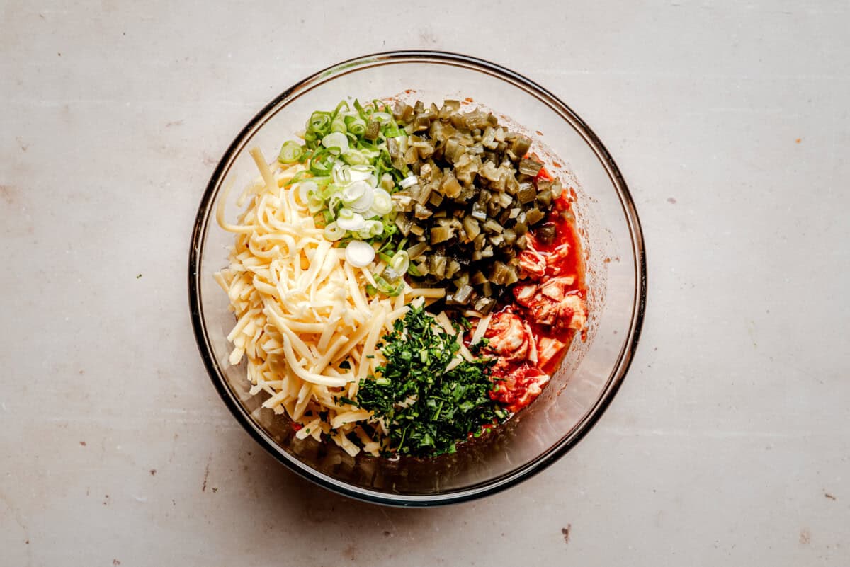 A glass bowl containing shredded cheese, sliced green onions, diced green chiles, diced cooked chicken, and chopped herbs, all arranged in separate sections on a light-colored surface.