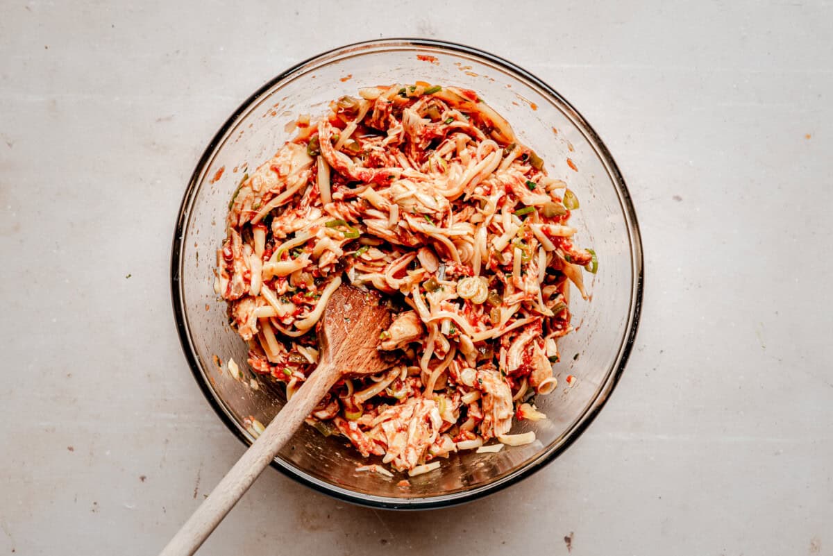 A glass bowl filled with shredded chicken and spices, being mixed with a wooden spoon.