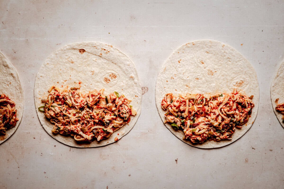 Four tortillas laid out on a light surface, each partially filled with a shredded chicken tinga, ready to be wrapped or rolled.