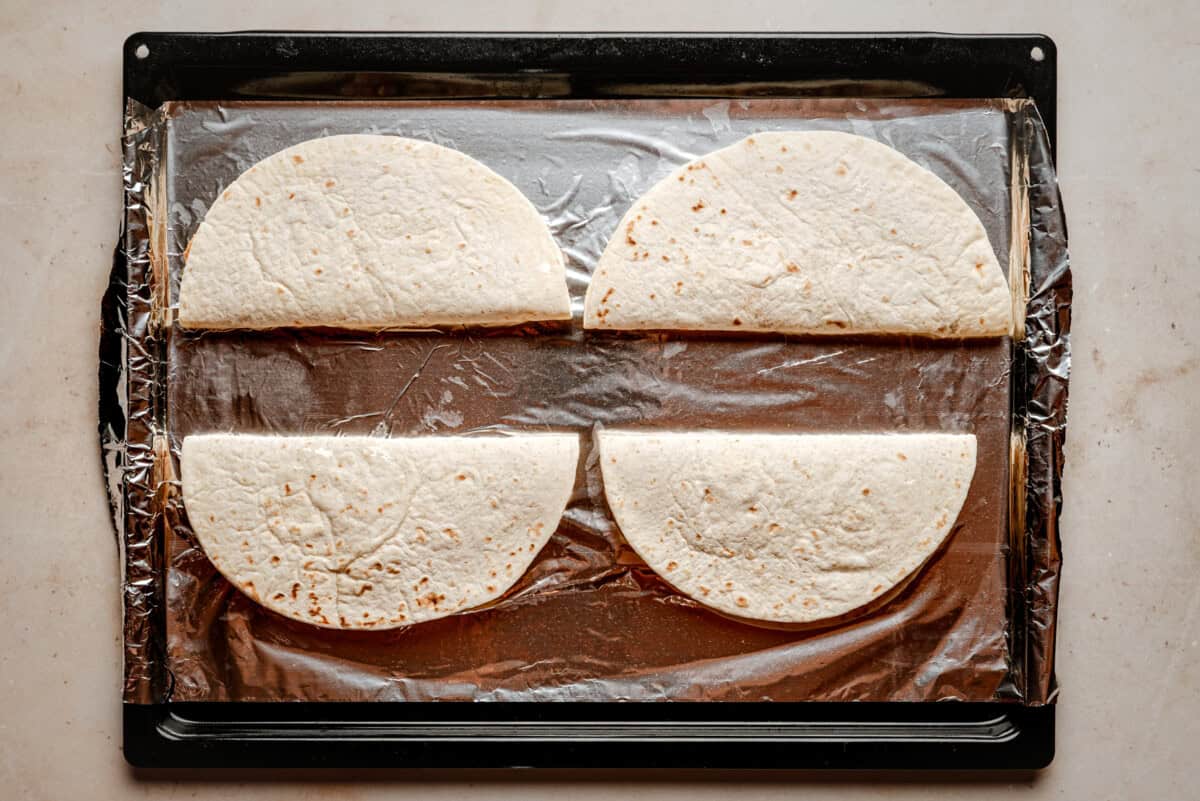 Four flour tortillas, each folded in half, are arranged on a baking sheet lined with aluminum foil. The baking sheet is placed on a light-colored countertop.
