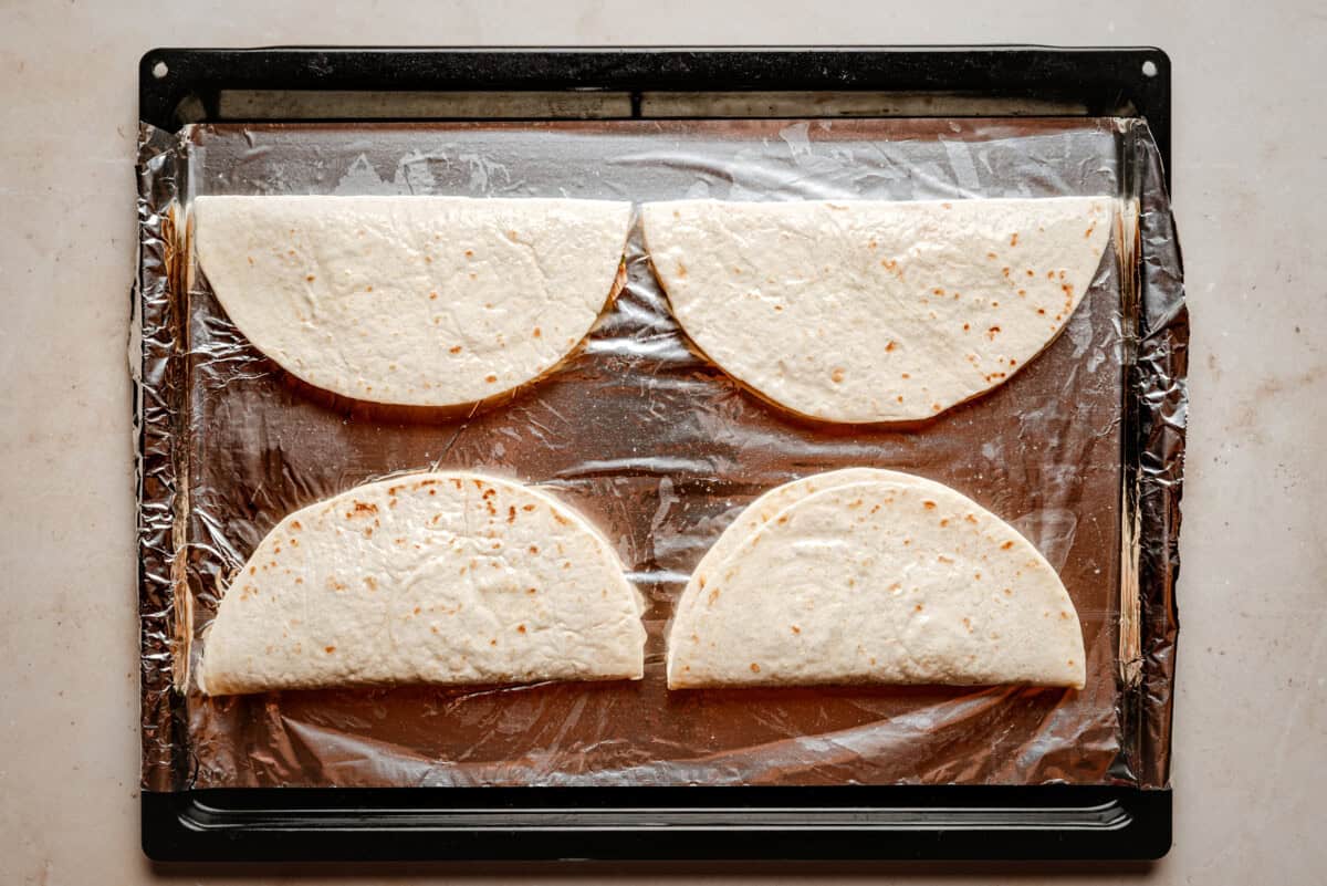 Four folded tortillas are arranged in a single layer on a baking sheet lined with aluminum foil, ready to be cooked.