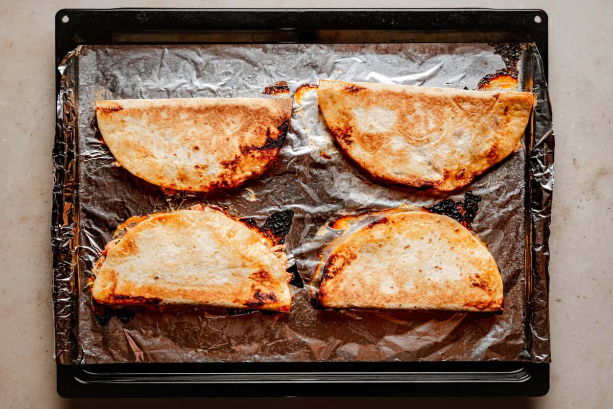 Four golden-brown chicken tinga quesadillas on a foil-lined baking tray, slightly crispy at the edges, resting on a light-colored countertop.