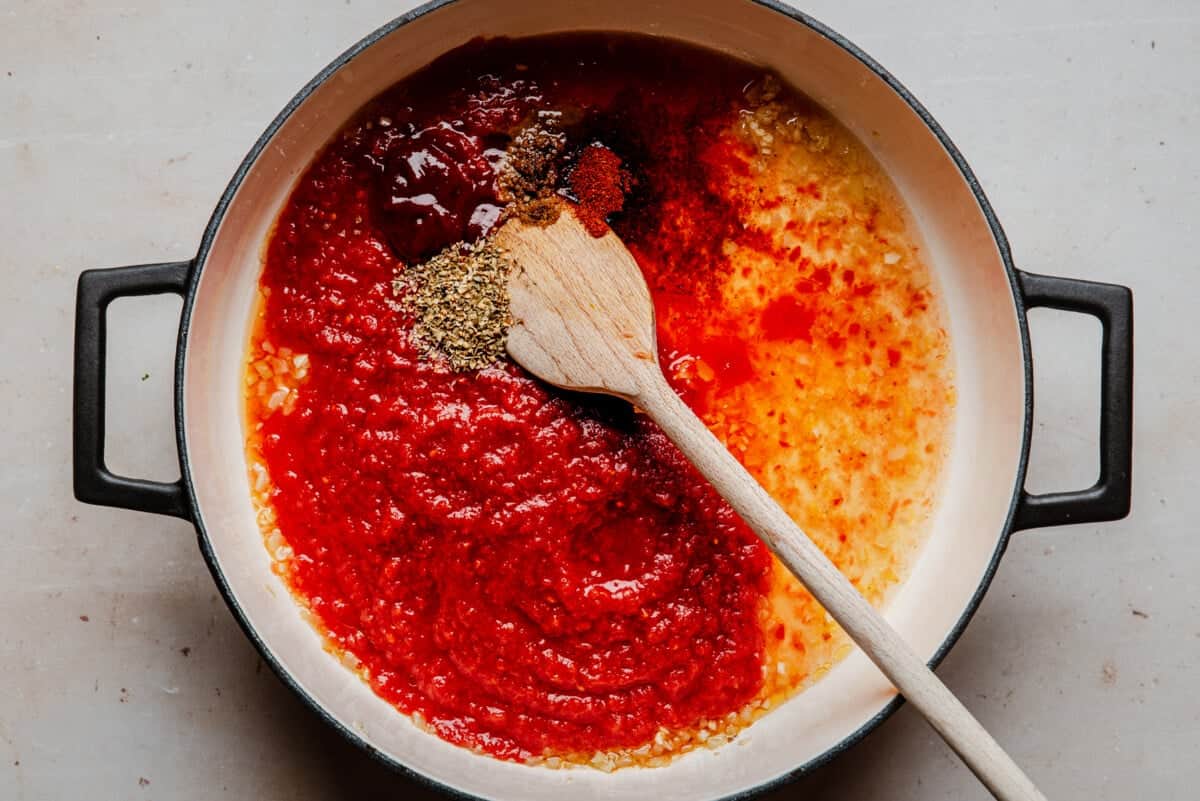 A pot with a wooden spoon contains tomato sauce, minced garlic, oil, and various spices, ready to be mixed together. The ingredients are grouped separately and have vibrant red and yellow colors.