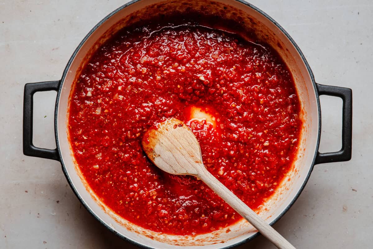 A pot of red tomato sauce simmering, with a wooden spoon resting inside, seen from above on a light-colored surface.