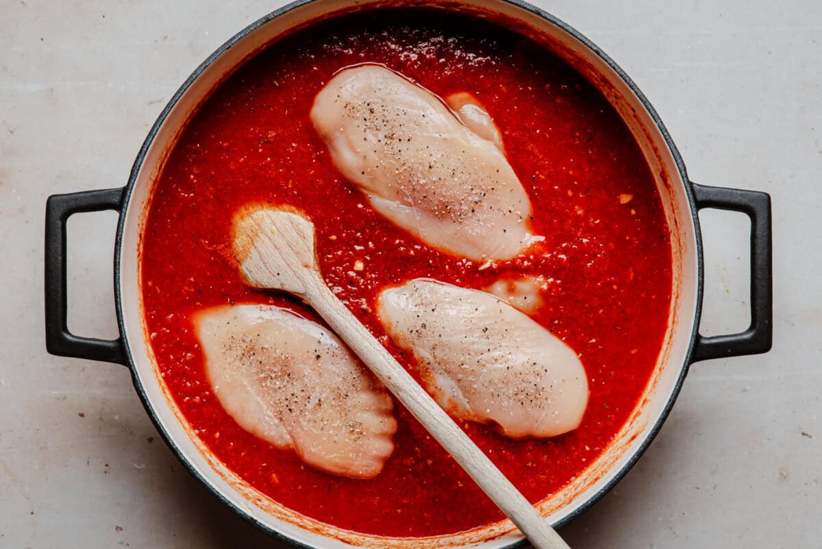 Raw chicken breasts sit in a pot of red tomato sauce, seasoned with pepper. A wooden spoon rests on the side, and the pot has black handles. The scene is viewed from above on a light surface.