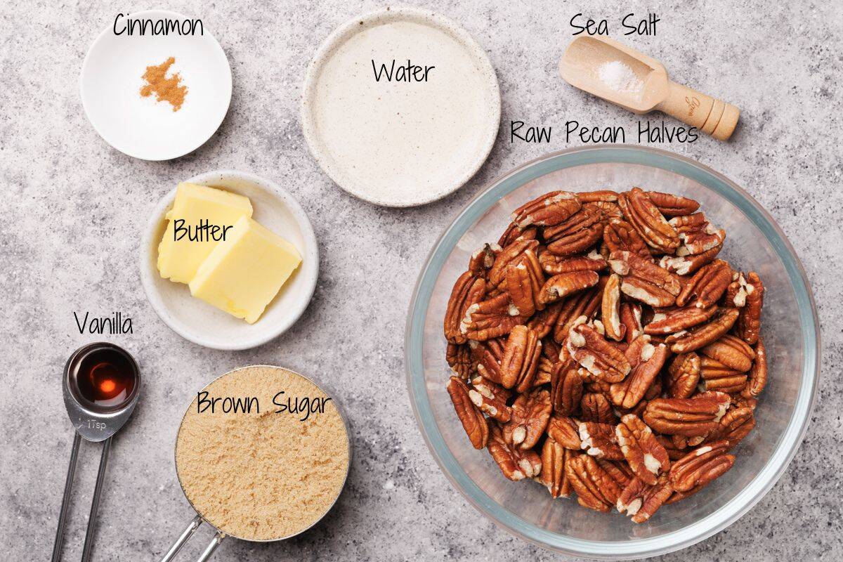 A top-down view of ingredients for praline pecans: a large bowl of raw pecan halves, a measuring cup of brown sugar, two butter slices, a spoon of vanilla, a dish of water, a pinch of cinnamon, and a scoop of sea salt.