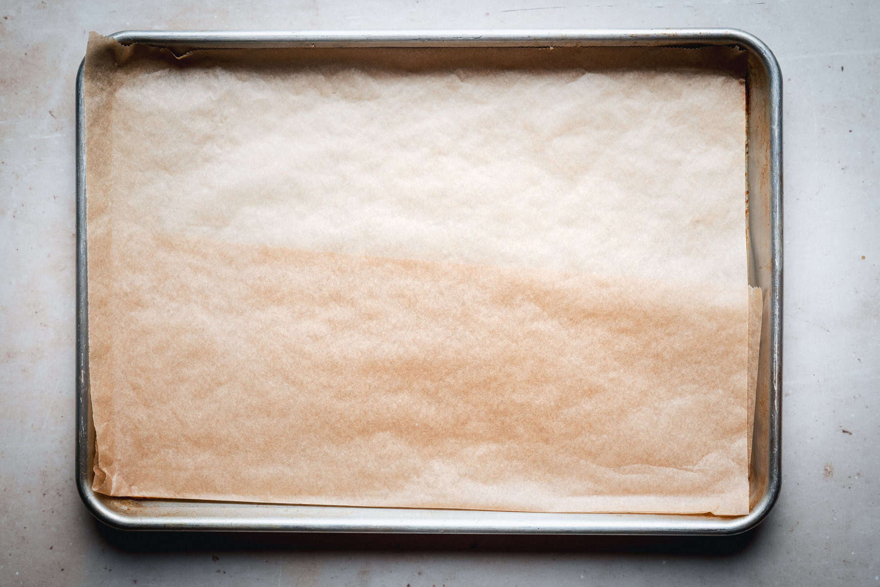 A metal baking sheet lined with a sheet of brown parchment paper, set on a light-colored surface. The parchment paper fits neatly inside the tray, ready for use in baking or roasting.