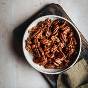 A bowl filled with praline pecans sits on a wooden board, with a brown cloth partially visible beside it on a light-colored surface.