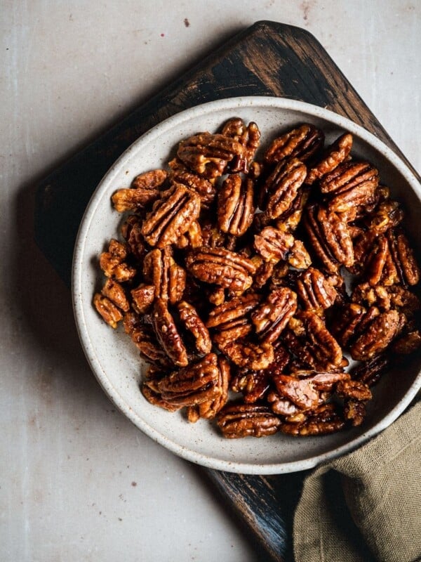 A bowl filled with praline pecans sits on a wooden board, with a brown cloth partially visible beside it on a light-colored surface.
