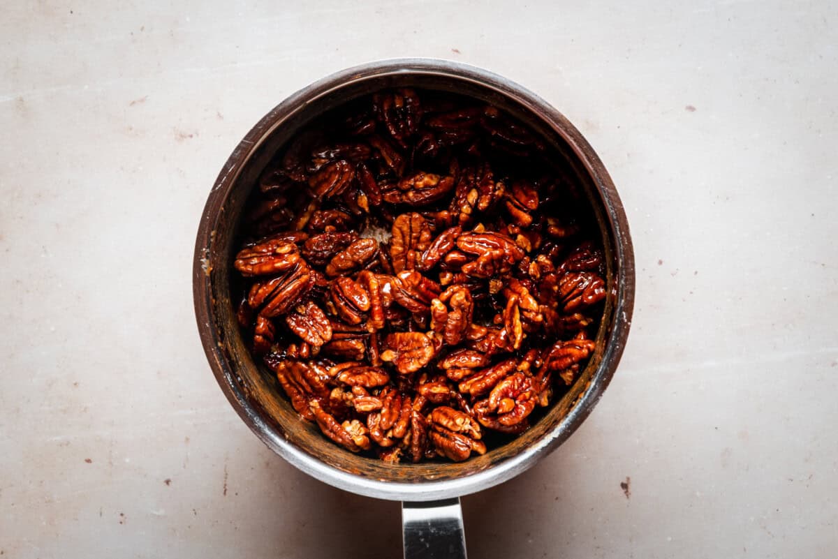 A metal saucepan filled with glazed pecans sits on a light beige surface. The pecans are shiny and coated, appearing roasted or caramelized.