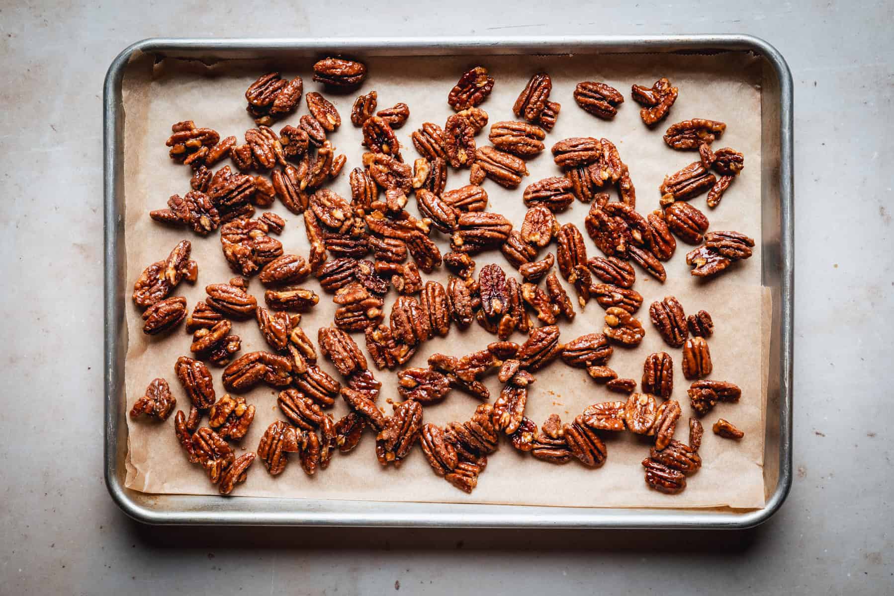 A baking tray lined with parchment paper holds an even layer of roasted, caramelized praline pecans. The nuts are golden brown and glistening, spaced out across the tray.