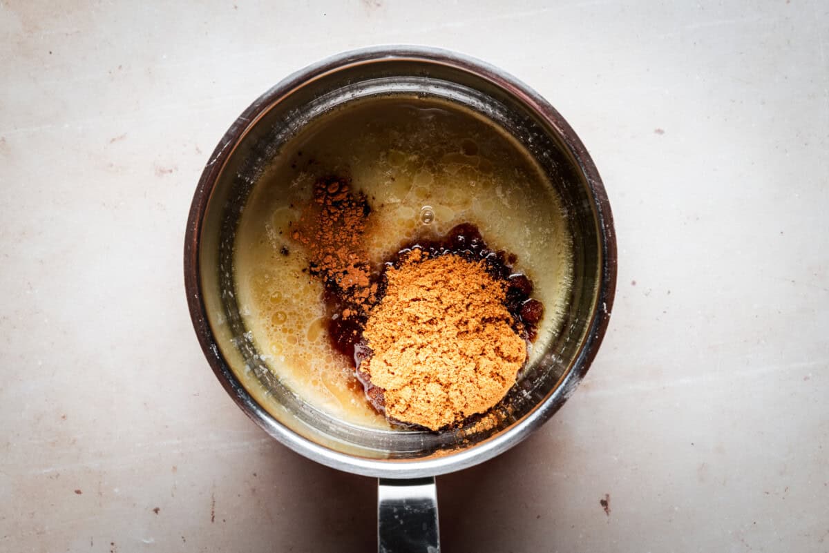 A saucepan viewed from above contains melted butter, brown sugar, and cinnamon, all partially mixed together, sitting on a light-colored countertop.
