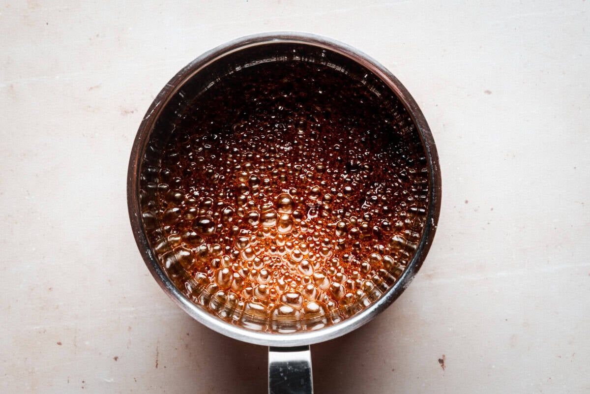 A metal saucepan filled with bubbling, golden-brown caramel sits on a light-colored surface, viewed from above.