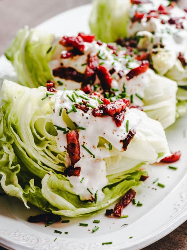 Three wedges of iceberg lettuce topped with creamy dressing, crispy bacon bits, and chopped chives are arranged on a white plate.