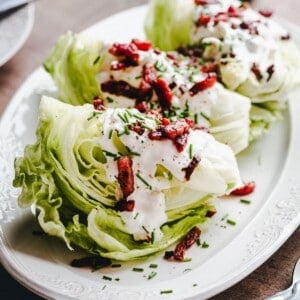 Three wedges of iceberg lettuce topped with creamy dressing, crispy bacon bits, and chopped chives are arranged on a white decorative plate.