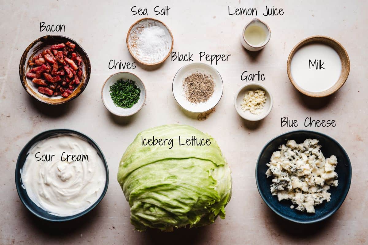 Ingredients for wedge salad arranged on a counter: bacon bits, sea salt, chives, black pepper, lemon juice, milk, sour cream, iceberg lettuce, minced garlic, and crumbled blue cheese, each labeled.