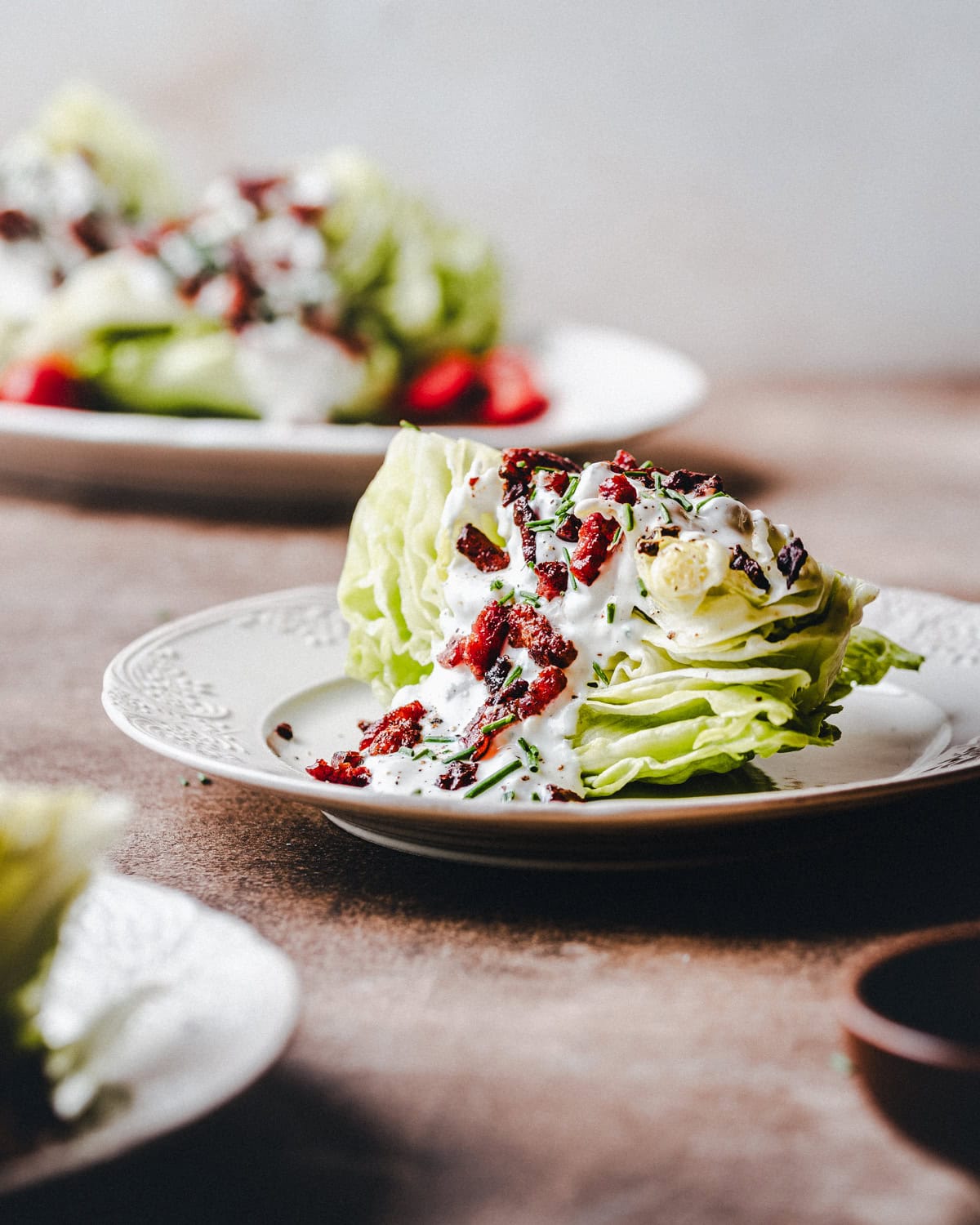 A wedge of iceberg lettuce topped with creamy dressing, chopped bacon, chives, and diced tomatoes is served on a decorative white plate. More salad plates are blurred in the background.