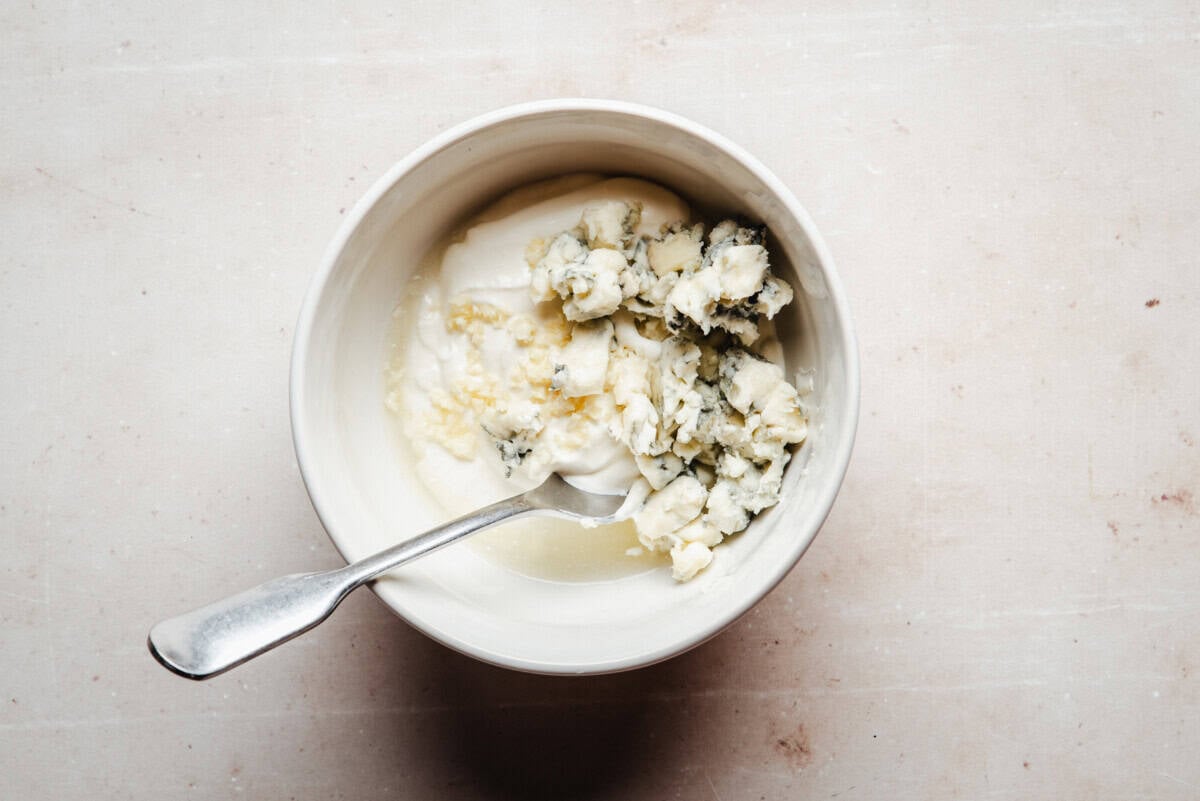 A white bowl containing sour cream and pieces of blue cheese, with a metal spoon resting inside, placed on a light-colored, textured surface.