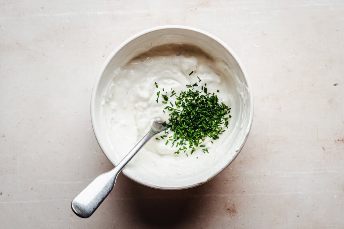 A white bowl filled with creamy white dip dressing with chopped chives, with a spoon resting inside the bowl, on a light-colored surface.
