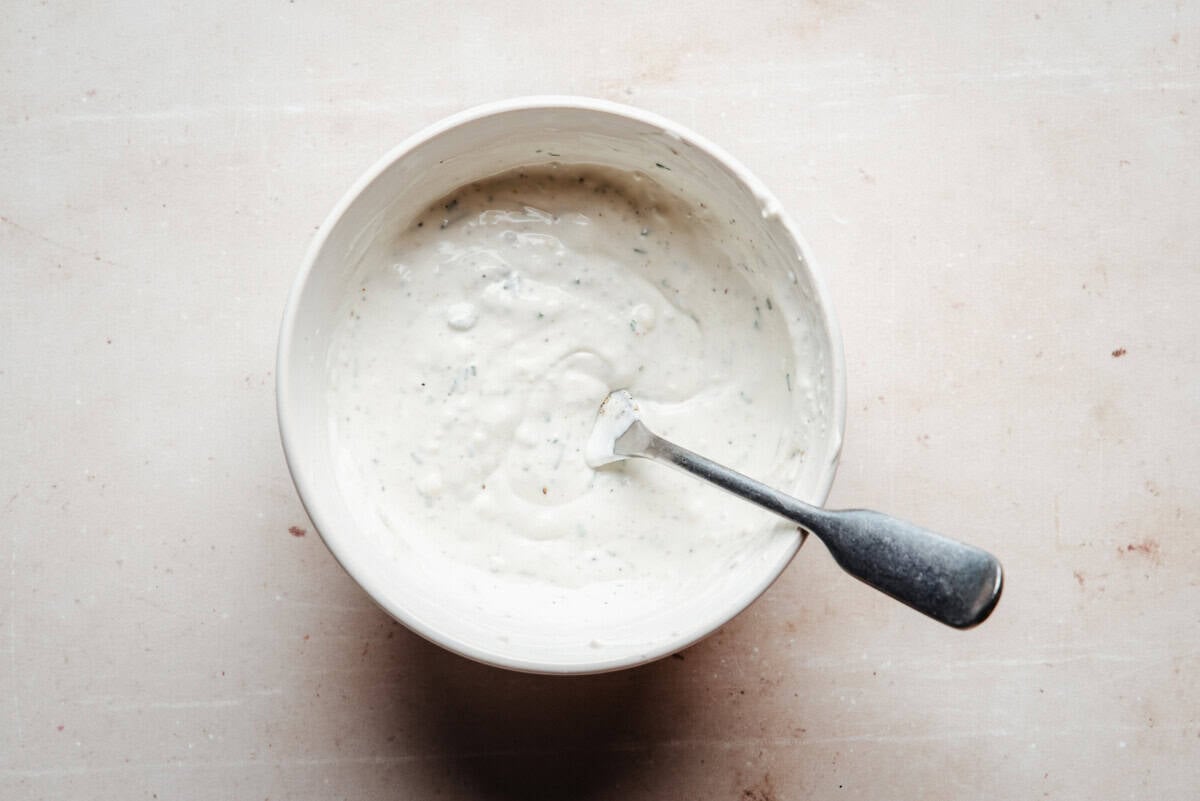 A bowl of creamy white dressing with herbs is shown on a light surface, with a metal spoon resting inside the bowl.
