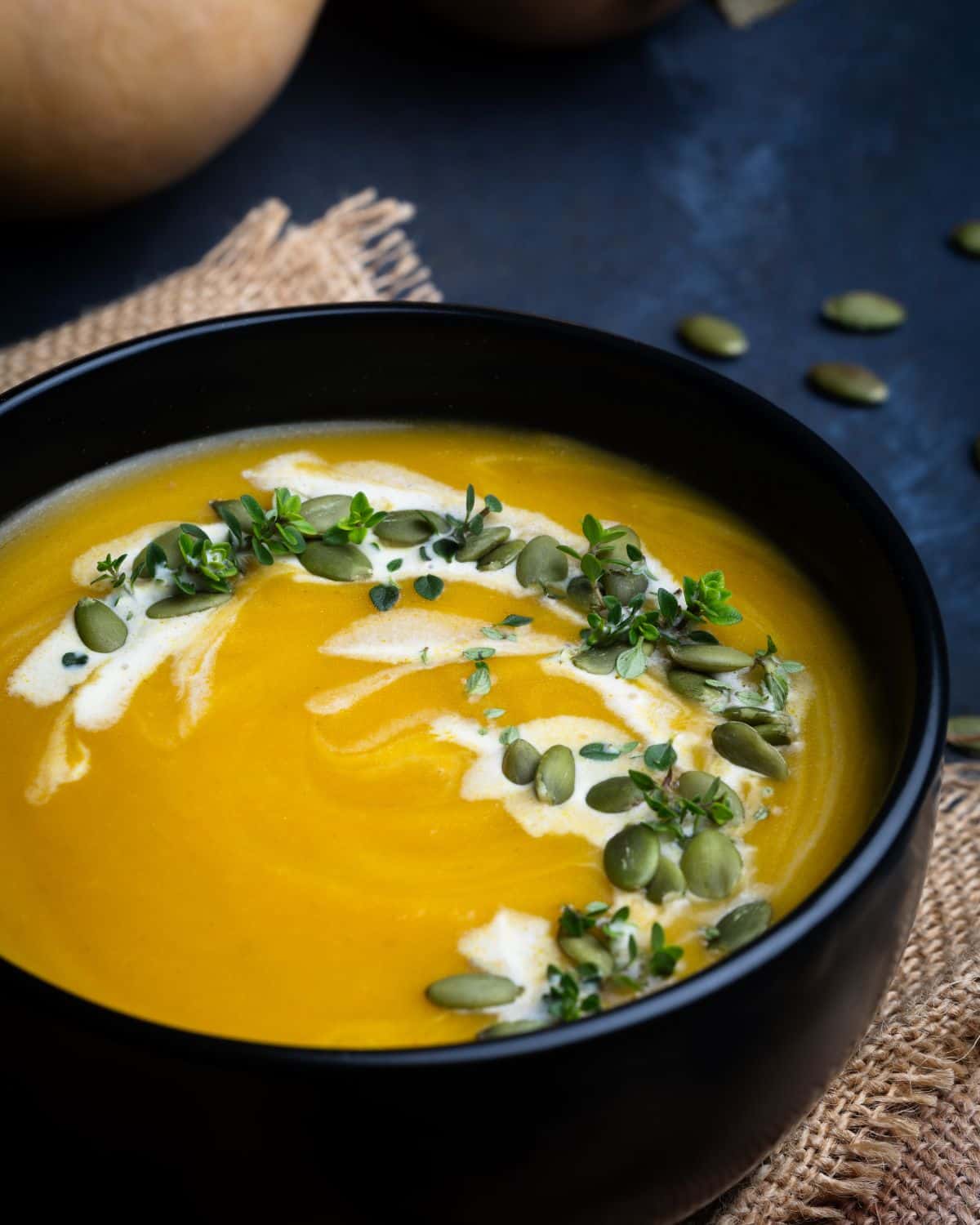 A bowl of creamy Butternut Squash Bisque garnished with a swirl of cream, fresh herbs, and pumpkin seeds, placed on a rustic burlap mat with scattered seeds nearby.