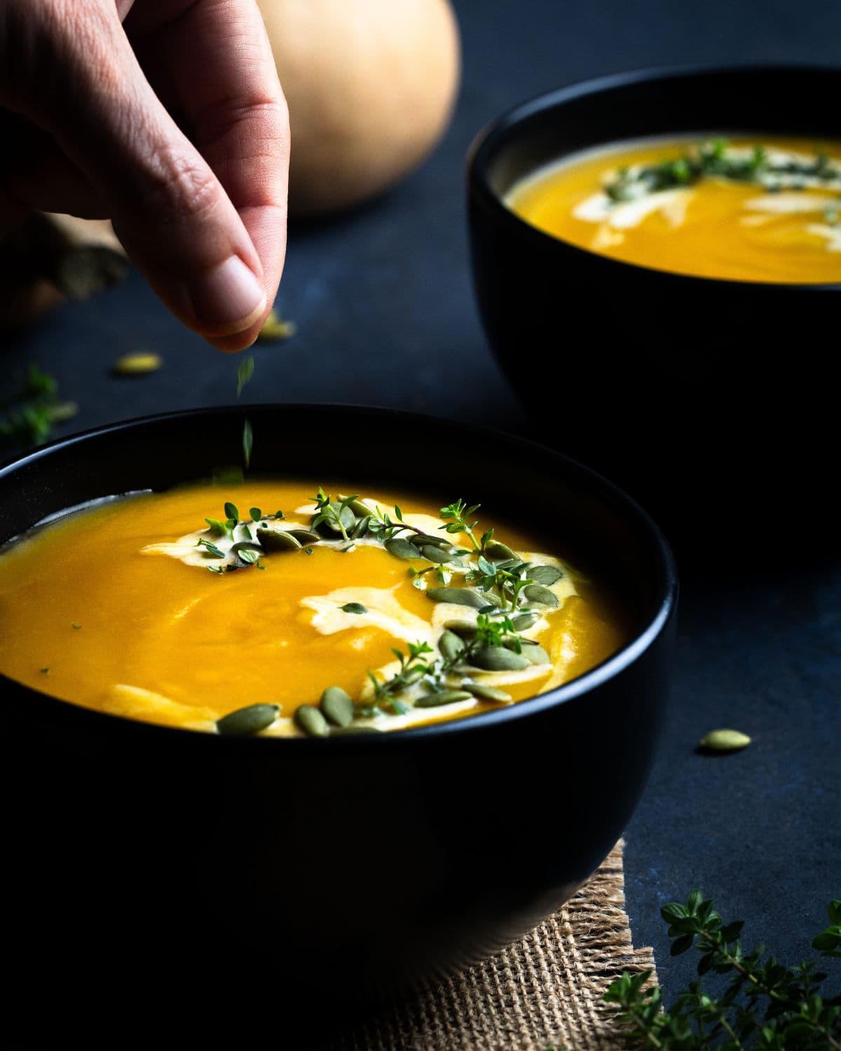 A hand sprinkles fresh herbs onto a bowl of Butternut Squash Bisque, garnished with pumpkin seeds and herbs, with another bowl of soup in the background on a dark surface.